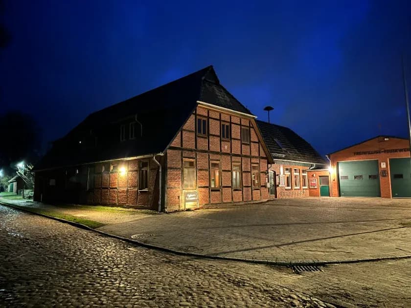 Das Kulturzentrum Witzeeze am Abend: ein rotes Fachwerkhaus mit warm erleuchteten Fenstern an der gepflasterten Dorfstraße, rechts daneben das Gerätehaus der Freiwilligen Feuerwehr